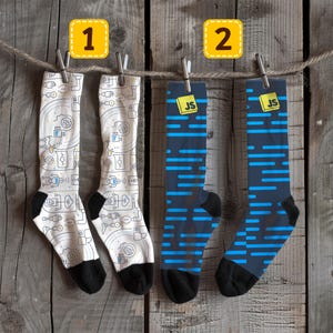 May include: Two pairs of knee-high socks hanging on a washing line. One pair is white with a repeating pattern of tech-related icons, black heels and toes. The other pair is navy blue with horizontal blue stripes, black heels and toes. Each sock has a yellow tag with the letters "JS".