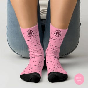 May include: Light pink socks with a black heel and toe. The socks feature a pattern of black binary code, the text "Hello World!", and a gear-like graphic. The socks are worn by a person wearing blue jeans.