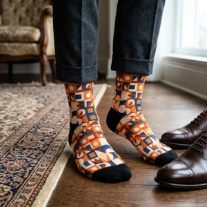 May include: A person wearing patterned socks with a geometric design in orange, blue, and white. The socks have black heels and toes. The person is wearing gray pants and brown leather shoes are visible on the floor.