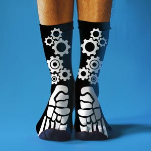 May include: Black socks with white gear and bone designs. The socks have a gear pattern on the upper part and a bone design on the foot. The socks are worn on legs against a blue background.