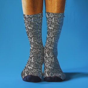 Può includere: Un paio di calzini grigio scuro con un motivo bianco a tema scientifico, tra cui becher, filamenti di DNA e molecole d'acqua. I calzini hanno talloni e dita blu navy, su uno sfondo blu brillante.