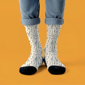Puede incluir: Un par de calcetines con un fondo crema y un patrón repetido de notas musicales negras, claves de sol y otros símbolos musicales. Los calcetines tienen talones y dedos negros. La persona lleva vaqueros azules.