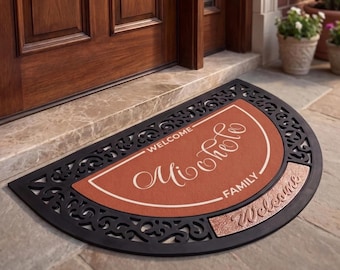 Op de afbeelding: Een halfronde deurmat met een zwarte decoratieve rand en een terracotta-kleurig midden. De mat bevat de witte tekst "WELCOME" en "FAMILY" rondom de in script geschreven naam "Michele", met "Welcome" in reliëf op de rand.