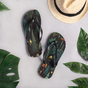 Könnte beinhalten: Schwarze Flip-Flops mit einem botanischen Design aus grünen Farnen, Pilzen und anderen Waldelementen. Die Flip-Flops haben schwarze Riemen. Das Bild zeigt einen Strohhut und grüne Blätter vor neutralem Hintergrund.
