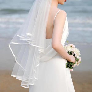 May include: A bride in a white wedding dress and veil, holding a bouquet of white and pink flowers. The veil has a ribbon trim. The background is a beach with the ocean visible.