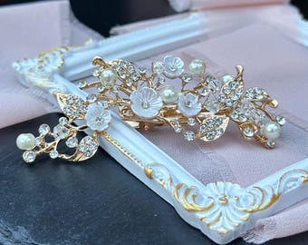 May include: Two gold hair clips with white flowers and rhinestones. The larger clip has a cluster of flowers and leaves, while the smaller clip has a single flower and leaves.