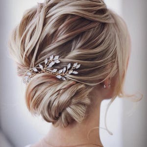 May include: A blonde updo hairstyle with a gold and crystal hair accessory. The hair is styled in a low bun with loose strands framing the face. The hair accessory is shaped like a branch with crystal leaves.