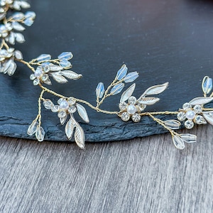 May include: A delicate gold-toned bridal headpiece featuring floral and leaf accents. The headpiece has clear crystal and pearl embellishments. The leaves are silver with a light blue hue. The headpiece is designed to be worn on the head.