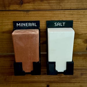 May include: Two black metal holders with rectangular blocks labelled "MINERAL" and "SALT". The mineral block is reddish-brown, and the salt block is white. The holders are mounted on a wooden surface.