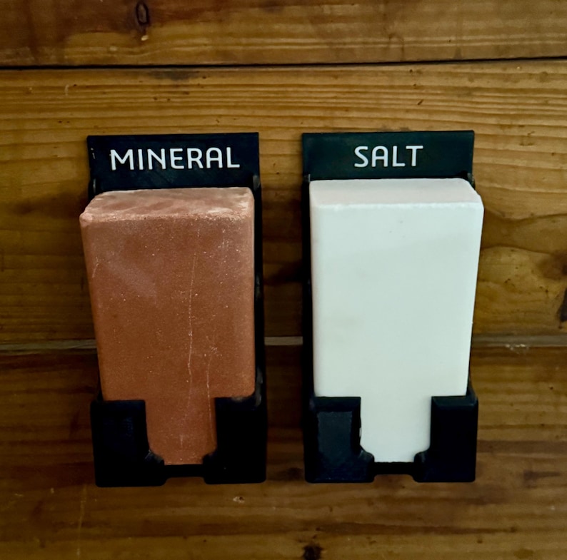 May include: Two black metal holders with rectangular blocks labelled "MINERAL" and "SALT". The mineral block is reddish-brown, and the salt block is white. The holders are mounted on a wooden surface.