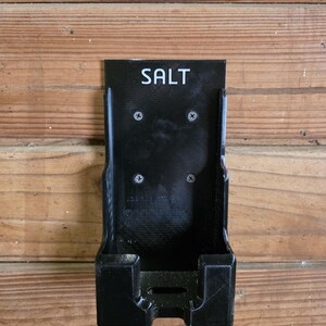 May include: A black, wall-mounted salt holder with the word "SALT" in white lettering. The holder is rectangular with a small shelf at the bottom and is mounted with four screws. The holder is made of a textured material.