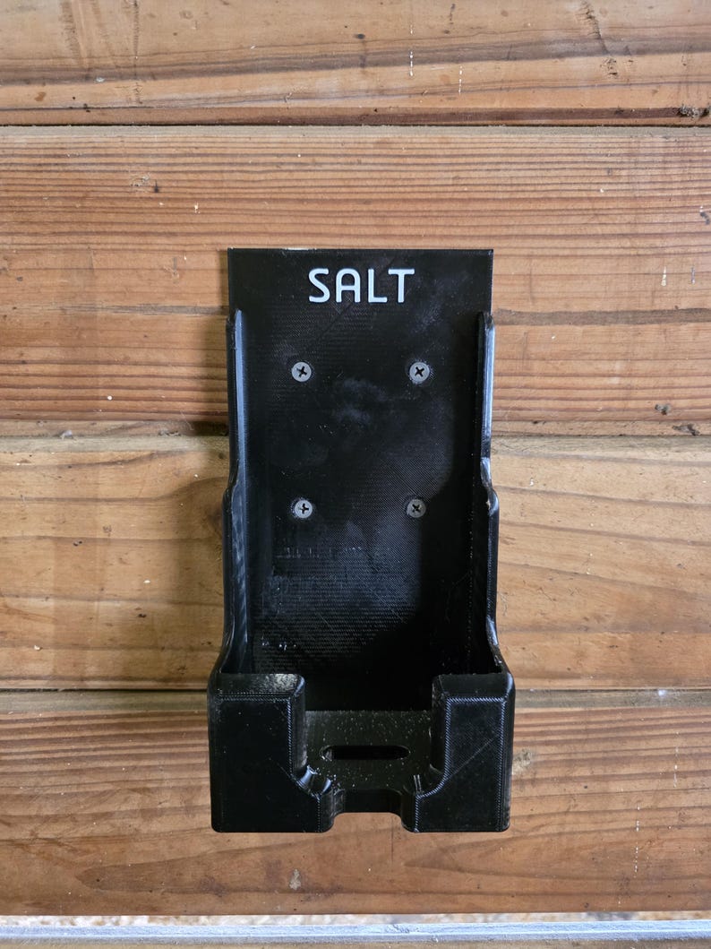 May include: A black, wall-mounted salt holder with the word "SALT" in white lettering. The holder is rectangular with a small shelf at the bottom and is mounted with four screws. The holder is made of a textured material.