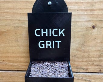 May include: A black, wall-mounted chick grit feeder with the words "CHICK GRIT" in white lettering. The feeder is filled with small, brown grit stones. The feeder is mounted on a wooden surface.