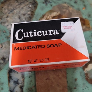 May include: A white box with a red and black label for Cuticura Medicated Soap. The label has the text "Cuticura" in black and "Medicated Soap" in white. The box has a net weight of 5.5 ounces.