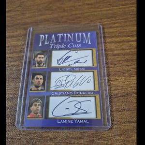 May include: A Platinum Triple Cuts sports trading card in a clear protective case. The card features portraits and ink signatures of Lionel Messi, Cristiano Ronaldo, and Lamine Yamal, with their names printed below each corresponding signature section.