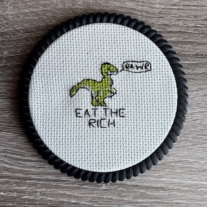 May include: A round, cross-stitched coaster with a black rope border. The center features a green dinosaur with a speech bubble saying "RAWR" and the text "EAT THE RICH" below.