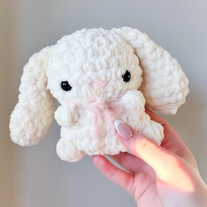 May include: A handmade, white crochet bunny plushie with long, floppy ears and a pink bow. The bunny has black eyes and a pink nose. The plush toy is soft and fluffy, perfect for a child's toy or nursery decor.