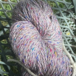 Peut inclure: Une pelote de laine de couleur lilas clair avec des reflets multicolores. La laine est enveloppée d'une ficelle blanche.