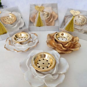 May include: A collection of flower-shaped incense burners featuring white or gold petals and a central gold-toned metal bowl with small perforations. Some burners are packaged in square boxes with a gold heart tag and a gold tassel attached.