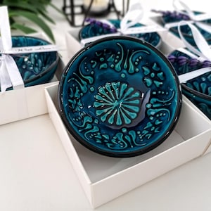 Puede incluir: Un cuenco pequeño y redondo de cerámica en color azul cerceta profundo con un patrón floral en relieve en un tono turquesa más claro. El cuenco está dentro de una caja de regalo blanca, atado con una cinta blanca y lavanda seca.