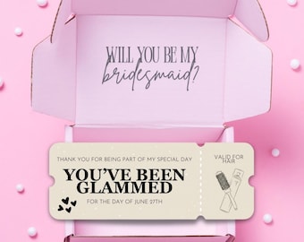 Może przedstawiać: Różowe pudełko z tekstem "Will you be my bridesmaid?" w środku. Pudełko zawiera bilet z tekstem "You've been glammed for the day of June 27th" i rysunkiem szczotki do włosów i lakieru do włosów.