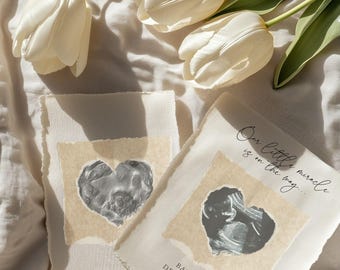 May include: Two cards with ultrasound images of a baby in a heart shape. The text on the card reads "Our little miracle is on the way... Baby Cook December 2026". The cards are on a white fabric background with white tulips.