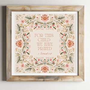 Op de afbeelding: Vierkant ingelijste kunstdruk met bloemenkransontwerp. De print bevat de tekst "FOR THIS CHILD WE HAVE PRAYED" en het bijbelvers 1 Samuel 1:27. Het kunstwerk heeft een neutrale achtergrond met perzik, roze en groene bloemelementen.