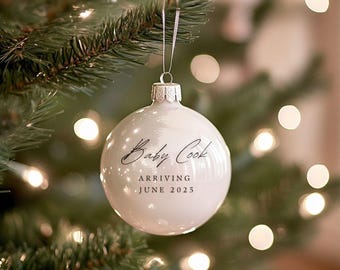 May include: White Christmas ornament with black text that reads "Baby Cook Arriving June 2025".