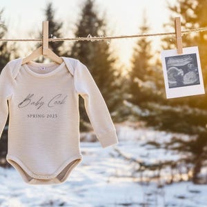 以下が含まれることがあります： 「Baby Cook SPRING 2025」の文字が入ったベージュ色のベビーボディスーツが、木製の洗濯バサミで洗濯紐に吊るされています。超音波写真がボディスーツの隣にクリップで留められています。背景には雪景色と常緑樹が写っています。
