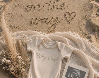以下が含まれることがあります： 砂の表面に「baby on the way」の文字とハートが描かれています。クリーム色のベビーロンパースに「Baby Cook」とプリントされ、超音波写真とドライフラワーも写っています。