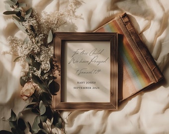 May include: A framed print with the text "For this child we have prayed 1 Samuel 1:27" and the text "Baby Jones September 2024". The print is on a white background and is framed in a rustic brown wood frame. The frame is sitting on a white fabric surface with a book and flowers.