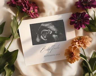 May include: A white card with a black and white ultrasound image of a baby. The card has the text "Baby [unclear text]" and "November 2024". The card is surrounded by pink and orange flowers.