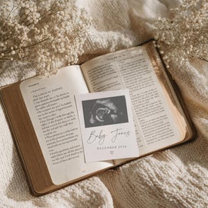 May include: An open book with aged pages, featuring text from Psalms, lies on a cream-colored blanket. A small card with a sonogram image and the text "Baby Jones December 2026" rests on the book. Delicate white flowers are scattered around the scene.