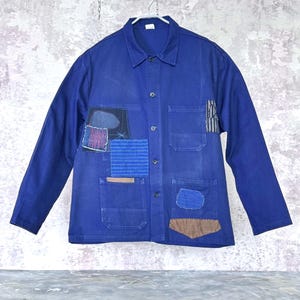 Puede incluir: Una chaqueta de trabajo azul real de manga larga con cuello clásico, cierre frontal de botones y varios bolsillos de parche. La prenda está decorada con varios parches de tela en tonos azules, marrones y estampados a rayas cosidos.