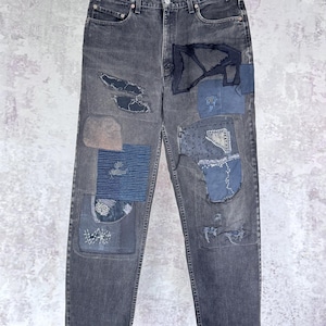 May include: Faded black denim jeans featuring a variety of stitched fabric patches in shades of blue and grey. The pants include visible mending, frayed edges, and decorative embroidery across the front legs, with a standard button fly closure.