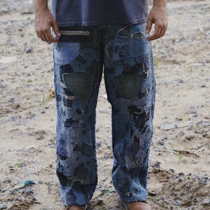 Può includere: Jeans in denim blu caratterizzati da un design patchwork molto usurato con molteplici tonalità di blu e nero, bordi sfilacciati, cuciture a vista e una tasca orizzontale con cerniera situata vicino alla cintura.