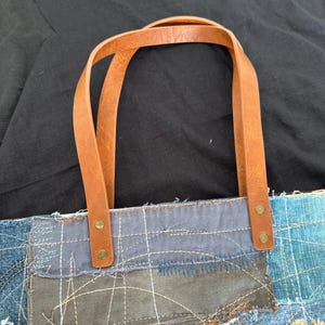 Pu&ograve; includere: Borsa tote in denim patchwork con manici in pelle marrone. La borsa presenta un mix di toppe in denim blu, cucite insieme con filo bianco visibile. I manici in pelle sono attaccati con rivetti color ottone.