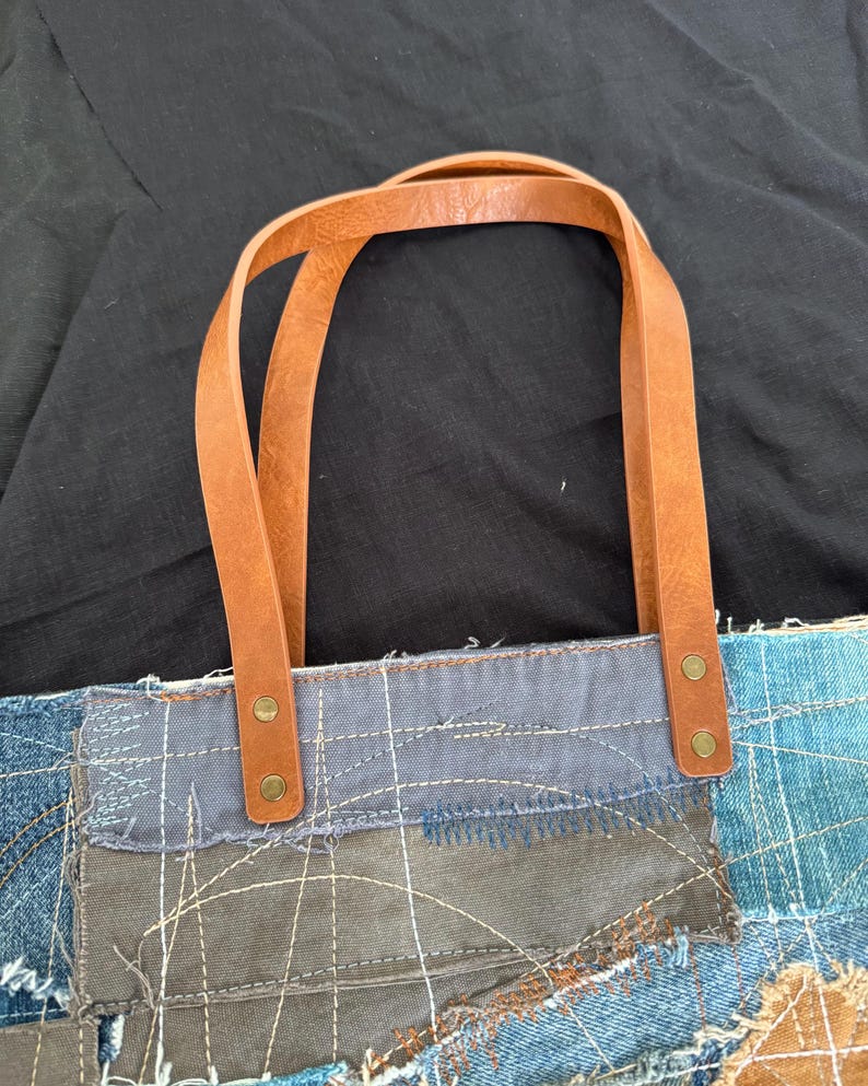 Pu&ograve; includere: Borsa tote in denim patchwork con manici in pelle marrone. La borsa presenta un mix di toppe in denim blu, cucite insieme con filo bianco visibile. I manici in pelle sono attaccati con rivetti color ottone.