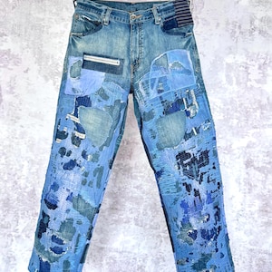 Può includere: Jeans in denim blu con un esteso patchwork e cuciture in stile sashiko in varie tonalità di blu. I pantaloni includono una tasca con cerniera sulla coscia destra, bordi in tessuto sfilacciato e un dettaglio in tessuto a righe sul retro.