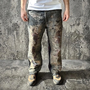 Könnte beinhalten: Lange Hose mit einem stark abgenutzten, mehrfarbigen Finish in Anthrazit-, Braun- und Cremetönen. Der Stoff weist eine raue, strukturierte Optik mit sichtbaren Ausfransungen und überlagerten Partien auf, kombiniert mit einem schlichten weißen T-Shirt.