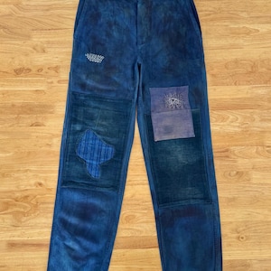 May include: Blue corduroy pants with patchwork details. The pants feature a button closure, pockets, and various patches in shades of blue. The patches include embroidered and textured designs, adding visual interest to the garment.