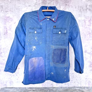 May include: A blue long-sleeve work shirt featuring a collar with red trim, two chest pockets with white buttons, and a small black label. The shirt displays white paint splatters, a large purple patch on the lower left, and a stitched square patch.