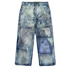 Può includere: Jeans in denim blu lavaggio acido con finitura usurata, toppe multiple, bordi sfilacciati e cuciture a vista. Il pantalone presenta una chiusura con bottoni, passanti per cintura e una silhouette a gamba larga in varie tonalità di blu e grigio.