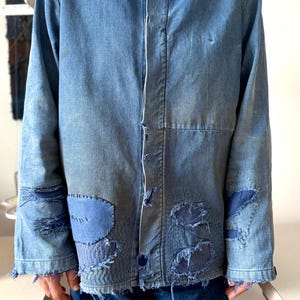 May include: A long-sleeve blue denim shirt featuring a collared neckline, a button-down front, and multiple distressed areas reinforced with darker blue fabric patches and visible stitching throughout the sleeves and lower front panels.