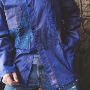 May include: A long-sleeved blue button-down shirt featuring multiple patches of varying blue fabric textures, including a frayed square patch and a vertically striped panel, worn over a grey undershirt with distressed blue denim jeans.