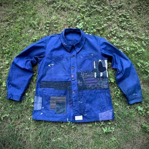 Könnte beinhalten: Eine blaue Arbeitsjacke mit mehreren Stoffflicken in verschiedenen Texturen und Mustern. Die Jacke hat eine Knopfleiste, einen Kragen und lange Ärmel. Farbspritzer sind sichtbar. Die Jacke liegt auf grünem Gras.