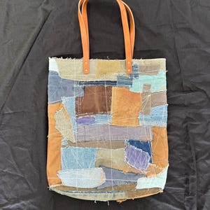 Pu&ograve; includere: Una borsa tote patchwork con toppe di tessuto in varie tonalit&agrave; di blu, marrone e beige. Ha due manici in pelle marrone fissati con rivetti in ottone. Una borsa unica e fatta a mano.