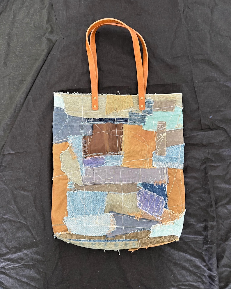 Pu&ograve; includere: Una borsa tote patchwork con toppe di tessuto in varie tonalit&agrave; di blu, marrone e beige. Ha due manici in pelle marrone fissati con rivetti in ottone. Una borsa unica e fatta a mano.