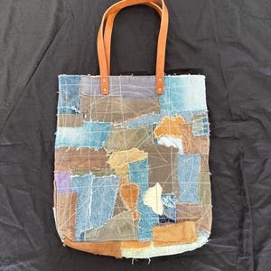 Pu&ograve; includere: Una borsa tote patchwork con toppe di denim in tonalit&agrave; blu, marrone e beige. Presenta manici in pelle marrone fissati con rivetti in ottone. La borsa ha una forma rettangolare con fondo arrotondato.