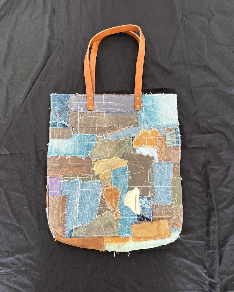 Pu&ograve; includere: Una borsa tote patchwork con toppe di denim in tonalit&agrave; blu, marrone e beige. Presenta manici in pelle marrone fissati con rivetti in ottone. La borsa ha una forma rettangolare con fondo arrotondato.